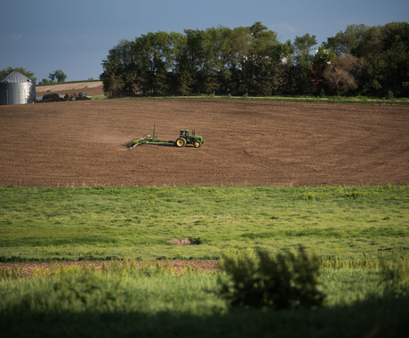 A tractor pulling a planter, planting in a bare field of brown soil. With a grassy area in the foreground and a tree line on the other side of the field.
