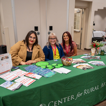 Tres mujeres detrás de una mesa con un mantel verde que dice Centro de Asuntos Rurales