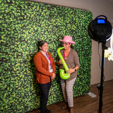 Two women at a photo booth, with a green leafy screen behind them