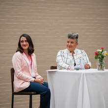 Two women sitting at a small table in front of a conference room, smiling at the audience