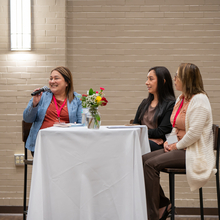 Three women at a small table, one woman holding a microphone