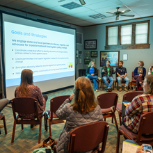 Panel of 3 females and 2 males speaks to an audience in a dark room with a projector screen