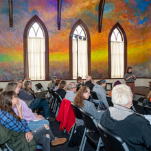 Several people sitting in a church with a mural painted on the ceiling. A woman stands at the front presenting. 