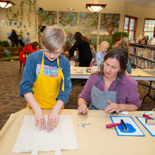 A child with glasses presses a stamp onto a cloth bag while a white female watches and assists him.