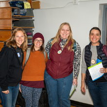 Four women smiling at the camera at the Local Foods Summit in a gym area with bleachers in the background.