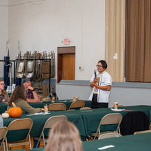 Hispanic male stands in a gym area presenting to several people sitting at tables. 