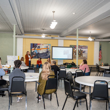 White female presenting in front of several people sitting around white tables. 