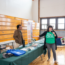 Black male stands behind a vendor table as a white female introduces him to the crowd