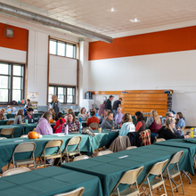 around 75 people sitting and standing in a gym conversing with each other.