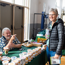 White male with grey hair sits at a vendor table and points to a while female with grey hair. They both are smiling at the camera.