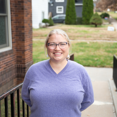 White female with blonde hair and glasses wearing a purple shirt stands in front of a brick building.