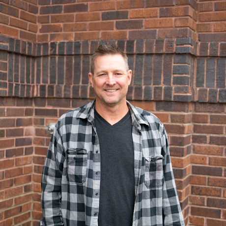 White male with brown hair wearing a black and grey plaid shirt stands in front of a brick building.