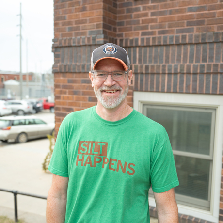 White male with grey beard, glasses, ball cap, and green shirt stands in front of a brick building