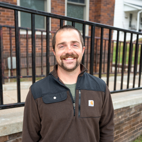 White male with dark hair and a beard and mustache stands in front of a black handrail of a brick building.