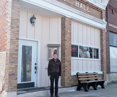 Woman with short dark hair, glasses, a dark jacket, and black pants smiles outside of a brick building labeled "CITY HALL."