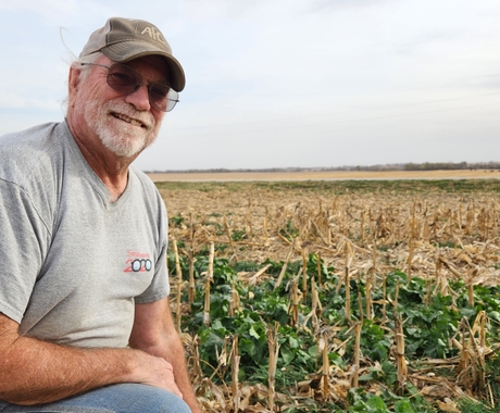 Man kneeling in field, with green plants growing among corn stalks. Man is wearing a gray Tshirt, ball cap, sunglasses, and has a white beard and mustache.