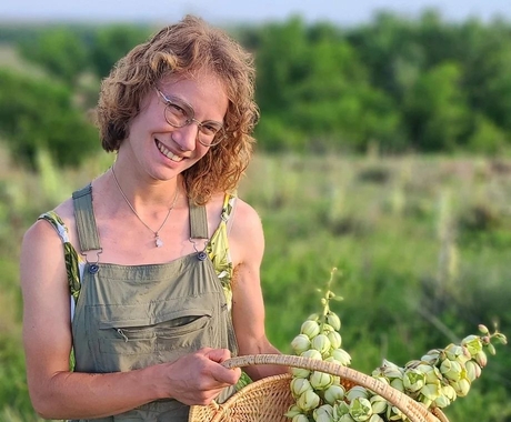 White woman with short, blond wavy hair, wearing glasses and green overalls holds a weaved basket with plants in it in a field of green plants behind her