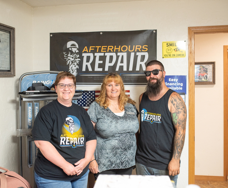Three people - a woman on the right and man on the left wearing "Afterhours Repair" black shirts, and a woman in the middle with a short sleeved gray shirt, standing in an office in front of a sign that says "Afterhours Repair"