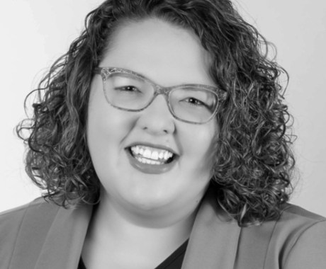 Mujer sonriendo a la cámara, con cabello castaño rizado hasta los hombros, gafas y una chaqueta sobre una camisa oscura.