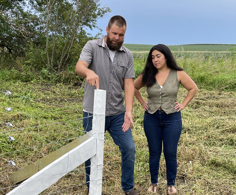 A man and woman stand in a field at a fence post corner. The fence post is made of a recycled wind turbine blade, and is white.