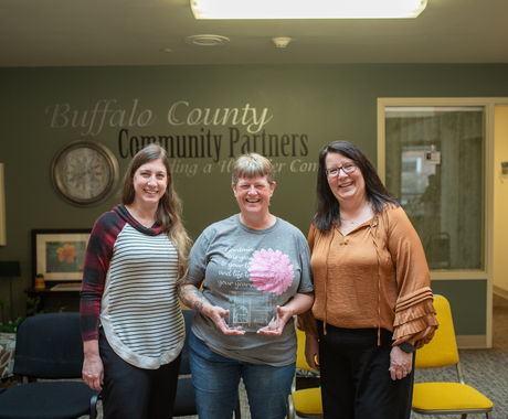 Three white females stand in a community room. The middle woman is holding an award plaque.