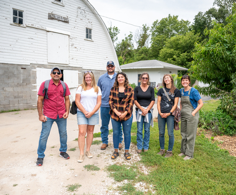 A group of 7 people standing in front of a white barn. The people range in age and gender and are wearing casual farm-visiting clothes like jeans and T-shirts.