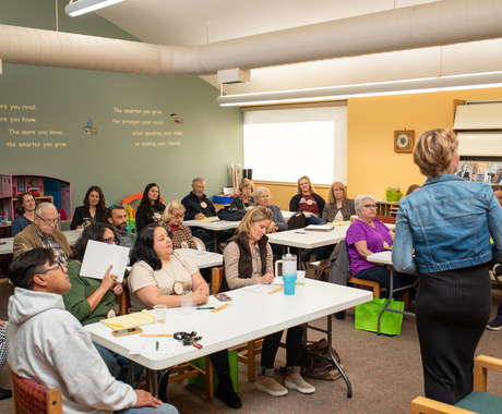 Group of more than 10 men and women sit at long tables facing towards a white woman with short blond hair wearing a jean jacket and black dress presenting to the group in a medium size room with green and yellow walls