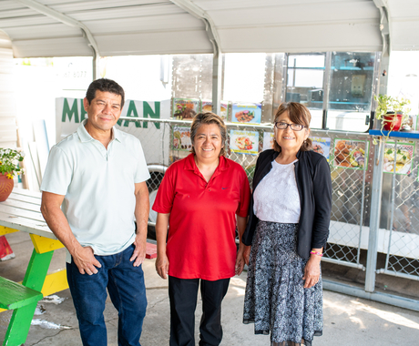 Three middle-aged looking people, man and two women, standing in front of their food truck and covered eating area, smiling at the camera.