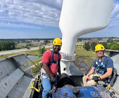 Two men wearing yellow hard hats sit on top of a wind turbine