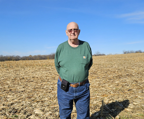 Man standing in a field covered in corn stalks. Man is wearing a green long-sleeved shirt, blue jeans, sunglasses, and has no hair.