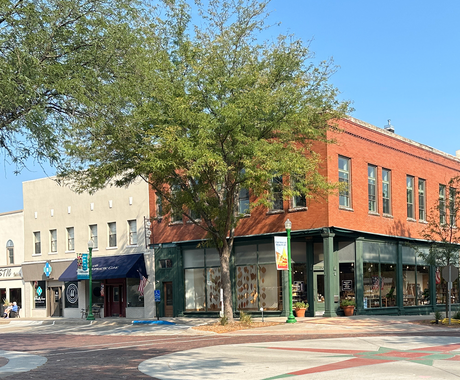 Main street corner in Ashland, NE. There is a tall, two-story red brick building with green posts on the corner with a two-story white brick building next to it. The red brick road has a cement circle at the intersection, with a red and green mosaic image in the center. 