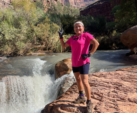 Indigenous woman wearing a pink shirt, black shorts, and black gloves, gives a peace sign while standing on red rock at the lip of a waterfall