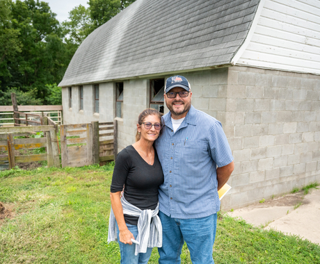 Man and woman standing in front of an old gray brick barn. Woman is wearing a black quarter sleeve shirt, glasses, and a light sweater tied around her waist. Man is wearing glasses, baseball cap, and a blue plaid button up shirt. 