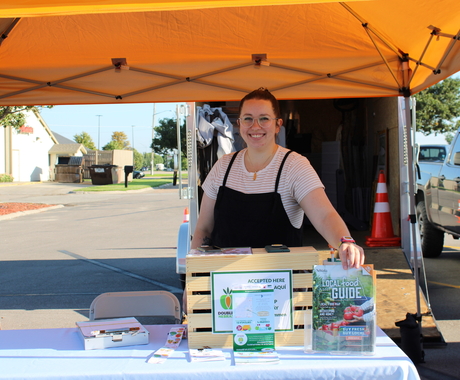 Una mujer de unos 20 años de pie detrás de una mesa y una exposición con alimentos locales. Está debajo de una carpa amarilla en un mercado de agricultores.