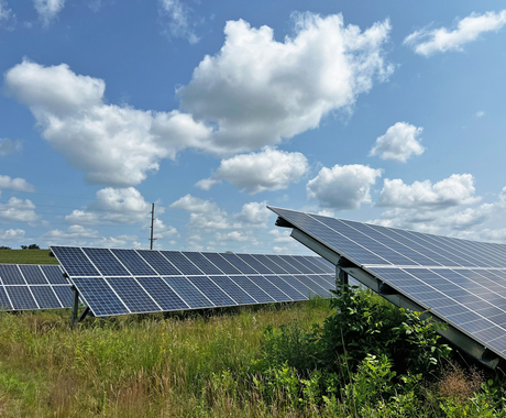 3 solar arrays with prairie grasses underneath. A blue sky with fluffy clouds above.