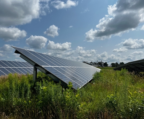 Two long solar arrays facing toward the right of the camera, with crops underneath. Sky is blue with fluffy clouds.