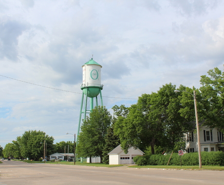A road leading into a community, with a white house and green and white water tower on the right.