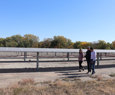 Three people walking in front of several rows of solar arrays. Green and orange trees in the background.