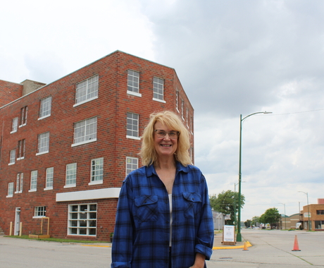 Woman wearing a blue plaid button up shirt, with glasses and blond shoulder-length hair, standing in front of a 4 story old building (a mill) with cloudy skies