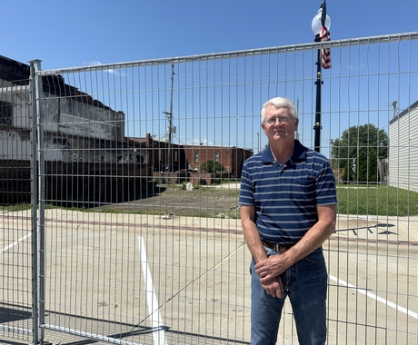 Man with gray hair wearing a blue polo with white stripes standing in front of a fence with an empty lot in the background