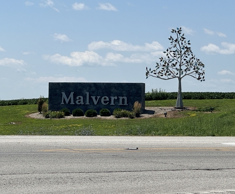 Sign at the corner of two highways, set on the ground with small ornamental plants around it. Says "Malvern." To the right of the sign is metal tree artwork.