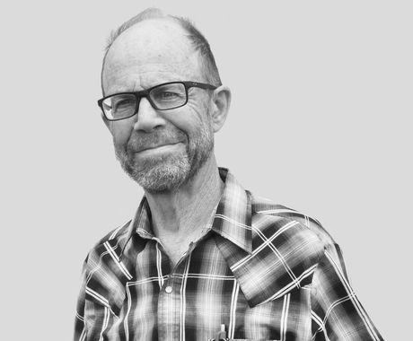 Man with balding hair, black large-rimmed glasses, wearing a dark plaid shirt, smiling at camera