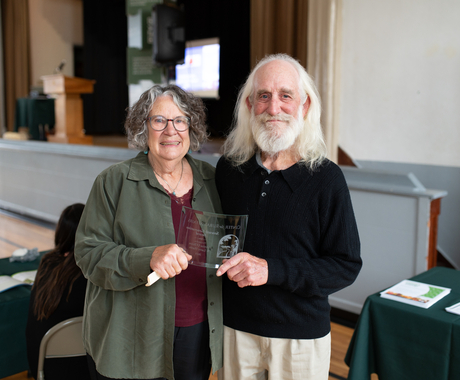 Woman and man holding a clear freestanding award plaque. Woman has short curly gray hair and is wearing a green long sleeve shirt. Man is wearing longer gray hair with a black long sleeved shirt.