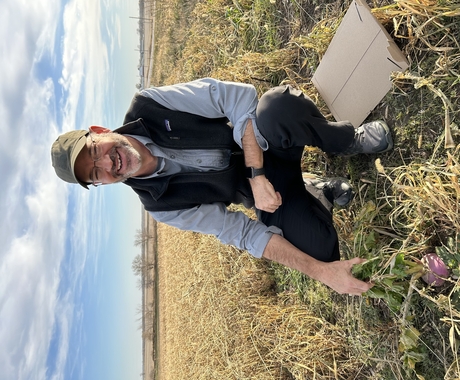 Man crouching in a field with a turnip at his feet. Man is smiling at the camera, wearing a green ball cap, glasses, long-sleeved button up shirt, black vest, and black pants.