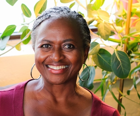 Mujer negra sonriendo directamente a la cámara. Lleva el pelo recogido hacia atrás, lleva unos grandes pendientes de aro, lleva una camiseta de color fucsia y hay plantas de fondo.