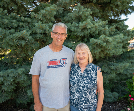 A man and woman in front of a tree, looking at the camera. The man has short white hair, glasses, and a gray T-shirt. The woman has shoudler-length blonde hair and is wearing a blue and white tank top.