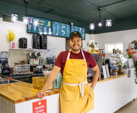 Man smiling at camera, leaning on a counter of a coffee shop. He's wearing a black ball cap, maroon short-sleeved shirt, and yellow apron