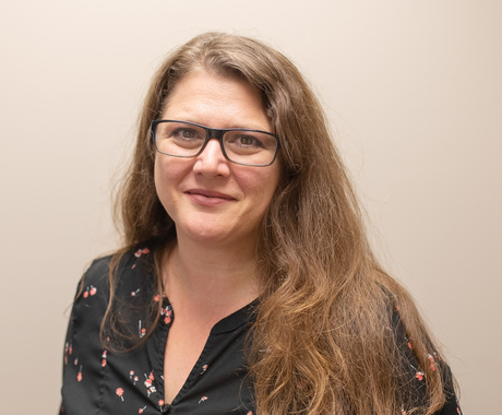 Woman wearing glasses looking at camera with a smile, with long, wavy, brown hair, wearing a black shirt with flowers