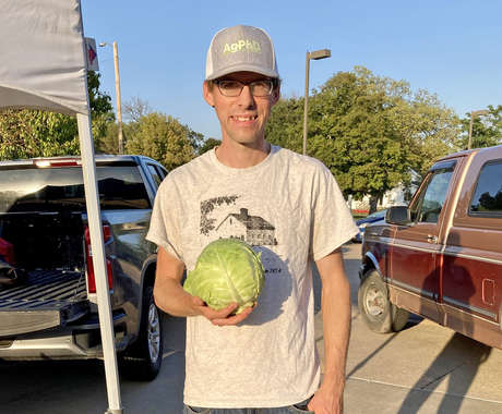 Man in ballcap, T-shirt, and jeans holding a lettuce, looking at the camera. In the background are the backs of 2 pick-up trucks and the leg of a pop-up tent.