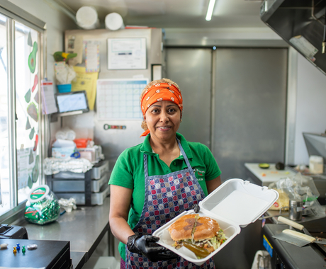 Latina woman wearing a red bandana, green shirt, and purple and pink checkered apron holds an open styrofoam container with a large sandwich inside. She's standing inside a food truck with the window to the left of the photo.